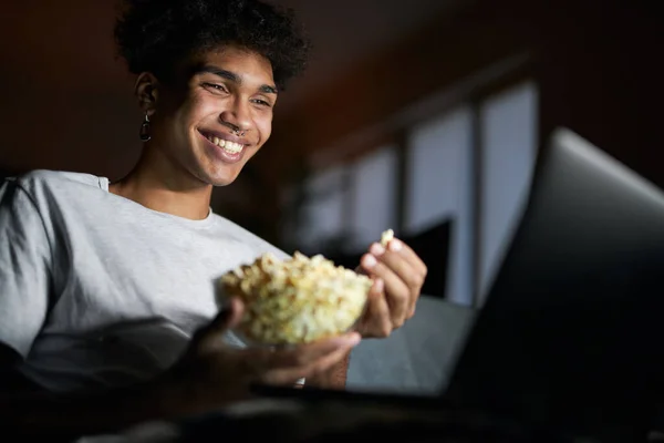 Šťastná sama. Radostný mladý muž se usmívá, sleduje komediální film na notebooku a jíst popcorn, zatímco sedí na pohovce v tmavé místnosti doma — Stock fotografie