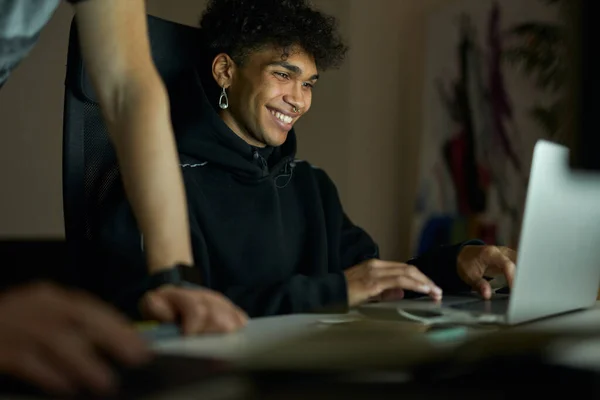 Love my job. Cheerful young guy with piercing smiling while sitting in front of laptop screen and working late in the evening
