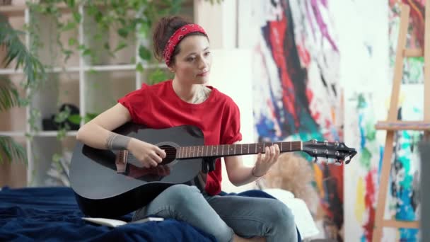 Вивчення нової навички. Зосереджена молода жінка грає на акустичній гітарі і слухає музику, сидячи на ліжку вдома у вітальні — стокове відео