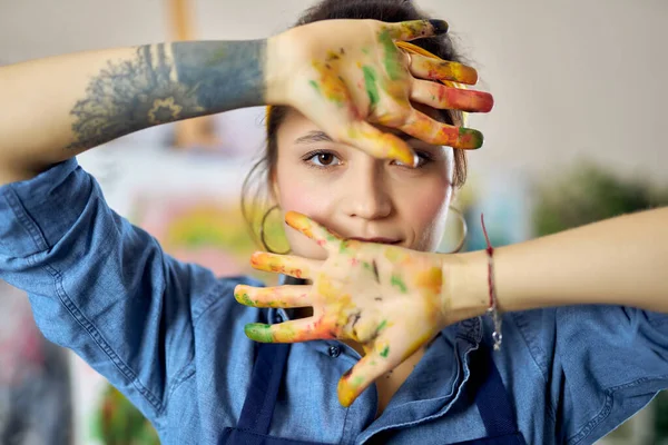 Potret wanita muda di celemek melihat ke kamera dan menunjukkan tangannya dalam cat saat melukis gambar di bengkel — Stok Foto