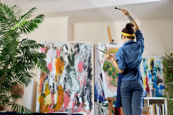 Pandangan kembali terhadap seniman perempuan mengangkat tangannya dengan kuas cat di dalamnya sambil menciptakan lukisan baru di atas kanvas, berdiri di bengkel studio rumah — Stok Foto