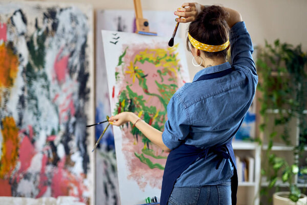 Back view of talented female artist holding paintbrushes with her hands in paint while thinking of a new painting, standing at home studio workshop