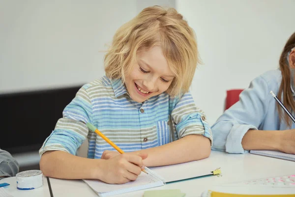 Vidám kis iskolás fiú mosolyog, ceruzát tart és jegyzetel, miközben tanul, ül az asztalnál az általános iskolai osztályteremben — Stock Fotó