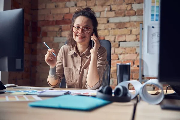 Creative woman, professional designer smiling at camera while making a call, working on new interior design project, sitting at the table in her office