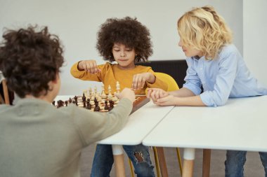 Afro saçlı harika bir çocuk. Diğer çocuklarla satranç oynarken konsantre olmuş görünüyor.