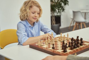 Mavi gömlekli akıllı çocuk sınıfta oturmuş satranç oynarken satranç tahtasına bakıyor.