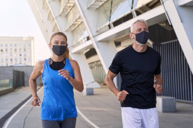 Korunaklı maskeler takan neşeli orta yaşlı çift Coronavirus salgını sırasında şehir ortamında birlikte koşuyor.