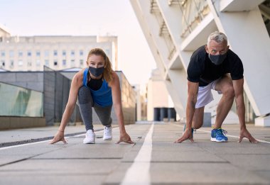 Başlangıç pozisyonunda koruyucu maskeler takan aktif olgun çift şehir ortamında birlikte koşmaya hazır.