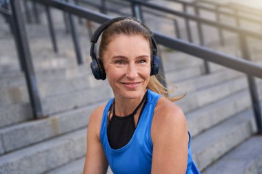 Spor kıyafetli, kulaklıklı, kameraya gülümseyen ve şehirde sabah antrenmanından sonra dışarıda poz veren orta yaşlı kadınların portresi.