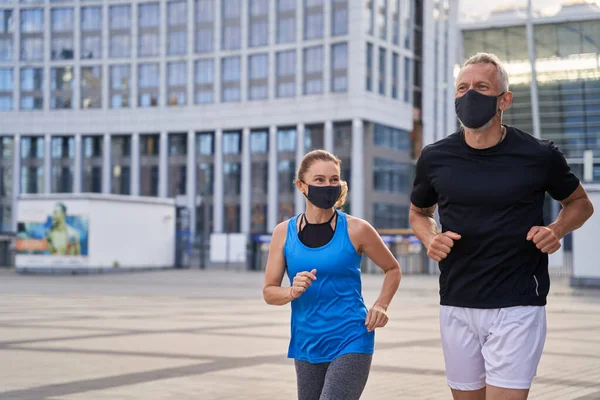Motivated middle aged couple, woman and man wearing protective masks jogging outdoors in the city to stay fit during Covid19 pandemic