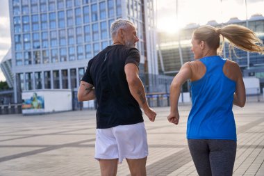 Spor giyim sektöründe orta yaşlı bir çiftin arka planda aktif kalıp şehir ortamında birlikte koşuşmalarının görüntüsü