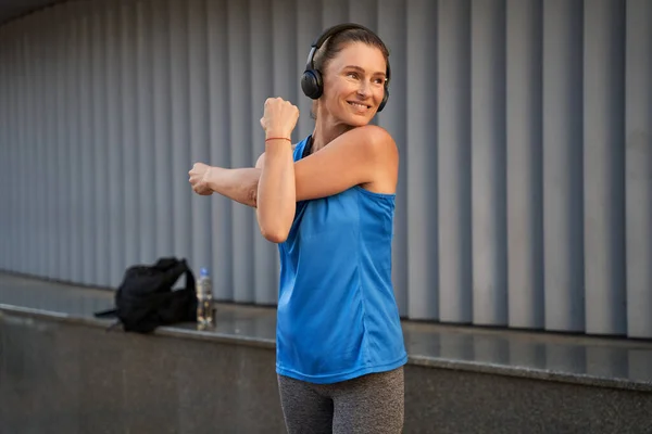 Mujer madura deportiva en ropa deportiva y auriculares sonriendo mientras se calienta, estirando los brazos, de pie al aire libre — Foto de Stock