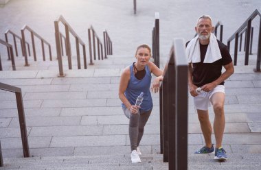 Aktif orta yaşlı çift, spor giyimli erkek ve kadın kameraya bakıyor, birlikte dışarıda egzersiz yaptıktan sonra merdivenlerden çıkıyorlar.
