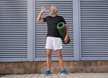 Sportif orta yaşlı adam su içiyor, spor paspası tutuyor, dışarıda spor yapmaya hazır.