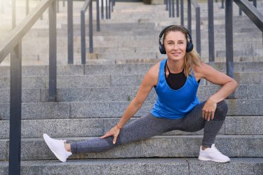 Neşeli orta yaşlı kulaklıklı kadın kameraya gülümserken bacaklarını açıyor, şehirde egzersiz yapıyor.