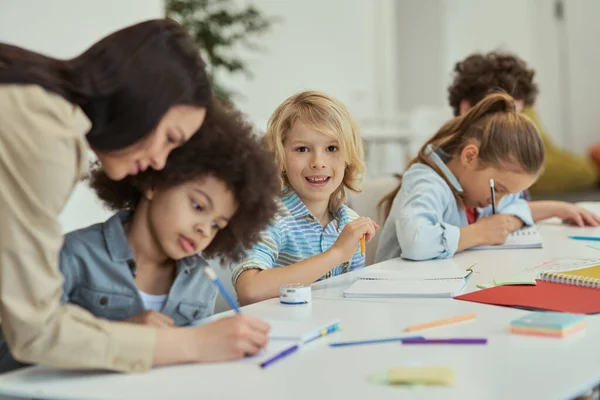 Avanzando. Pequeño colegial sonriente mirando la cámara mientras está sentado en la mesa de clases de la escuela primaria junto con otros niños — Foto de Stock