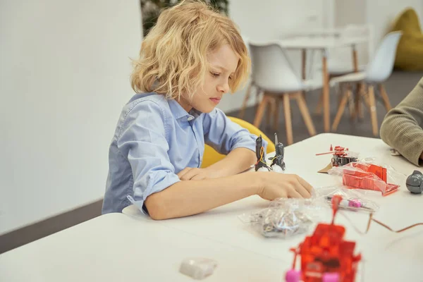 Menino caucasiano concentrado sentado à mesa e examinando detalhes de um brinquedo técnico durante a aula STEM — Fotografia de Stock