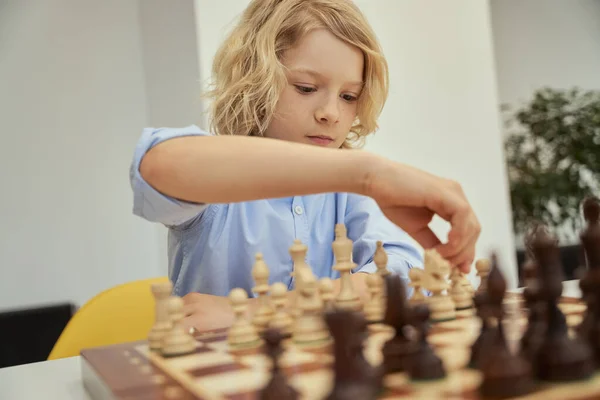 Портрет умного белого мальчика в синей рубашке, сидящего в классе и делающего ход во время игры в шахматы на шахматной доске — стоковое фото