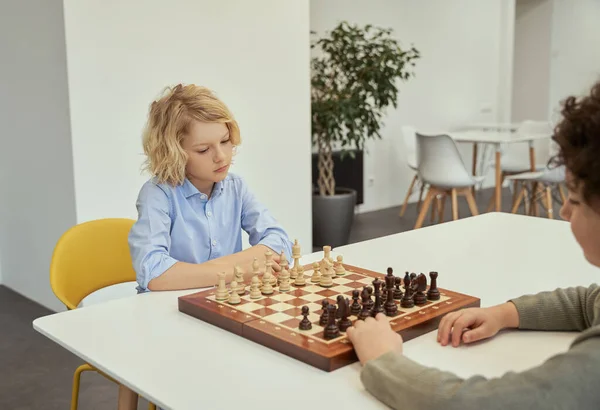 Думай, прежде чем действовать. Задумчивый маленький кавказский мальчик играет в шахматы со своим другом, сидя вместе за столом в школе — стоковое фото