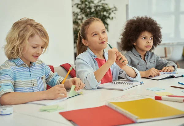 La educación es clave. Encantadores niños diversos estudiando y tomando notas mientras se sientan juntos en la mesa en el aula de la escuela primaria — Foto de Stock