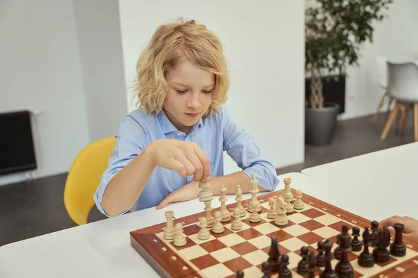 Сосредоточенный кавказский мальчик в синей рубашке сидит в классе и планирует свой ход, играя в шахматы на шахматной доске — стоковое фото