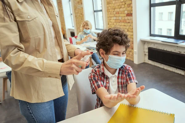 Niño de la escuela con máscara protectora listo para limpiarse las manos. Profesora usando un spray de alcohol para desinfectar las manos de los estudiantes en el aula — Foto de Stock