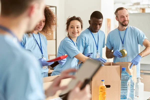 Vrolijke diverse vrijwilligers glimlachen terwijl ze voedsel sorteren in kartonnen dozen, en werken samen aan een donatieproject. Focus op de vrouw — Stockfoto