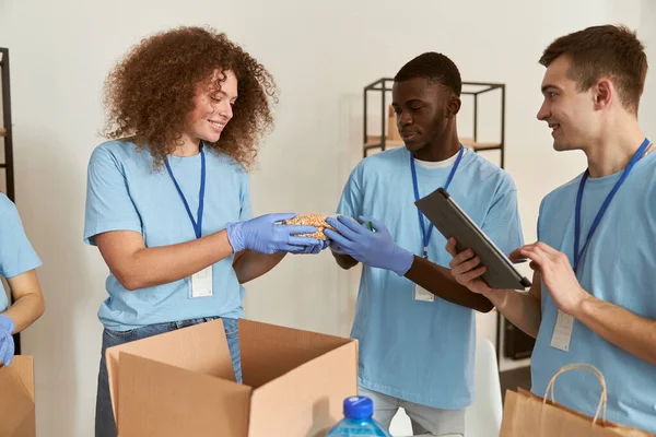 Команда молодых добровольцев в защитных перчатках улыбается во время сортировки, упаковывает продукты питания в картонную коробку, работает вместе над проектом пожертвования в помещении — стоковое фото