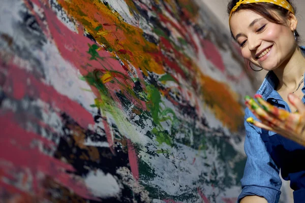 Pelukis wanita muda yang ceria menciptakan lukisan minyak abstrak modern yang besar, menerapkan cat di atas kanvas dengan jari-jari di bengkel — Stok Foto
