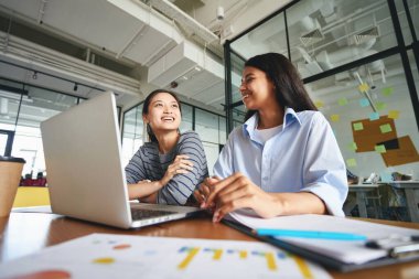 Başarılı genç bayanlar dizüstü bilgisayarları ve kağıtlarıyla masada oturuyorlar.