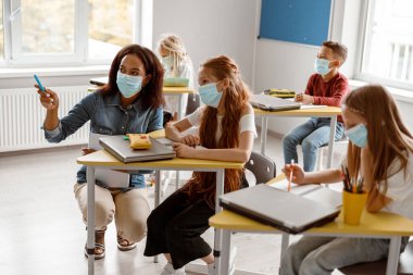 Bayan öğretmen kız masasının yanında çömeliyor.