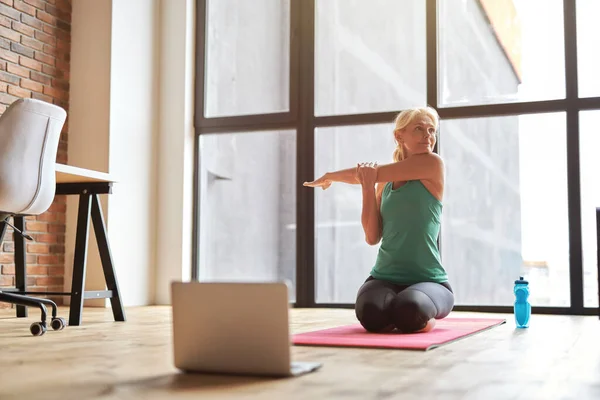 Обаятельная зрелая блондинка смотрит в сторону, сидя на полу и практикуя йогу с ноутбуком дома днем — стоковое фото