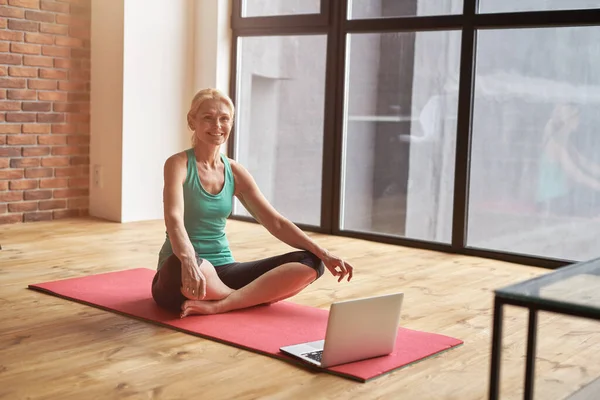 Urocza, dojrzała kobieta siedząca w Lotusie, pozuje na podłodze i uśmiecha się medytując i ćwicząc jogę w domu, po zajęciach online na laptopie — Zdjęcie stockowe