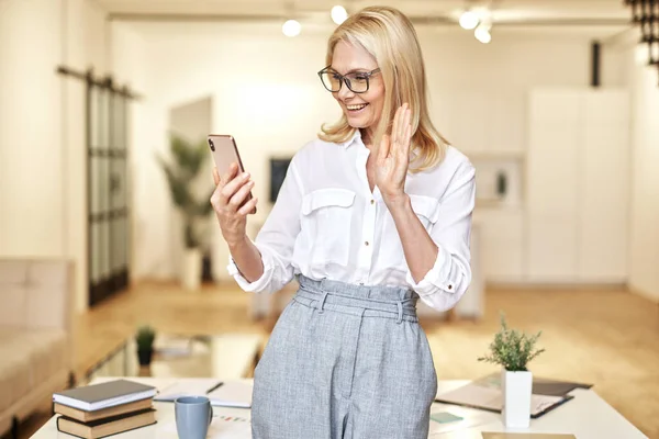 Pengusaha wanita dewasa yang bahagia melambaikan tangan pada kamera, memiliki panggilan video online menggunakan smartphone saat berdiri di kantor — Stok Foto