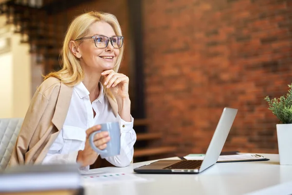 Pengusaha wanita dewasa yang ceria dengan kacamata tersenyum, menggunakan laptop dan minum kopi saat bekerja di kantor Stok Gambar
