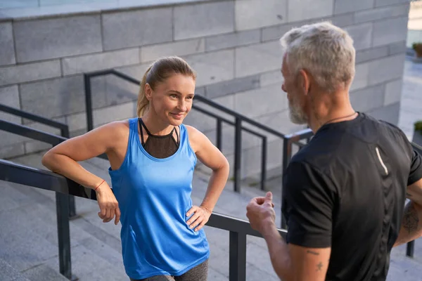Piękna sportowiec uśmiechnięta podczas rozmowy z mężem lub przyjacielem, stojąc na świeżym powietrzu po treningu w mieście — Zdjęcie stockowe
