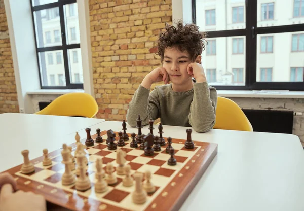 Наблюдать, как маленький мальчик сидит в классе и играет в шахматы на шахматной доске — стоковое фото