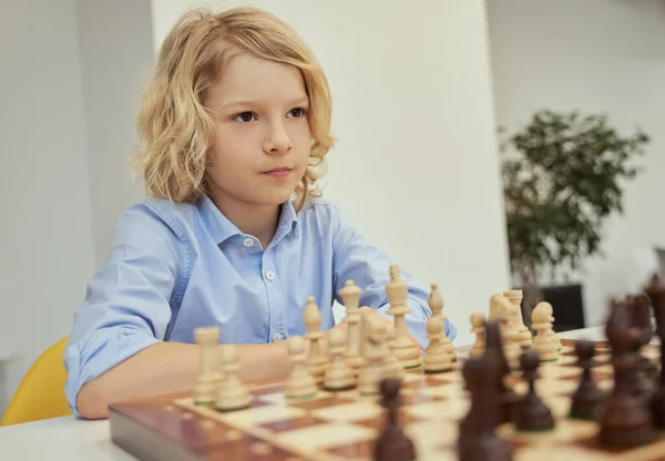 Портрет спокойного белого мальчика в синей рубашке, сидящего в классе и смотрящего в сторону, играя в шахматы на шахматной доске — стоковое фото