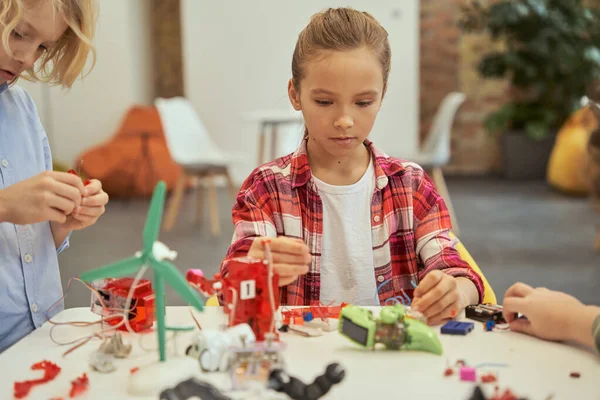 Entusiasta menina llittle olhar concentrado ao colocar um kit de brinquedo eletrônico juntos, sentado à mesa durante a aula STEM — Fotografia de Stock