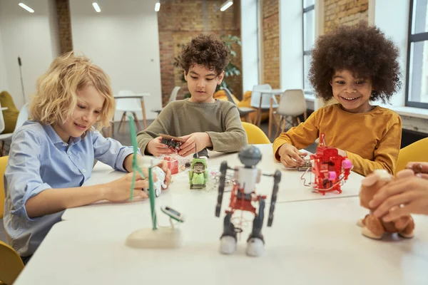 Інновації. Цікаві маленькі різноманітні хлопчики посміхаються, сидячи за столом і перевіряючи іграшки роботів, проводячи час разом в інженерному клубі — стокове фото