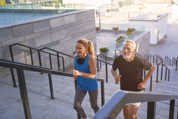 Mature couple in sportswear running up a flight of stairs in city while exercising together outdoors. Active sport, fitness, healthy lifestyle of middle aged people