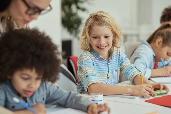 Aktiv liten skolpojke ler och tittar på sin klasskamrat när han sitter vid bordet i grundskolans klassrum tillsammans med andra barn — Stockfoto