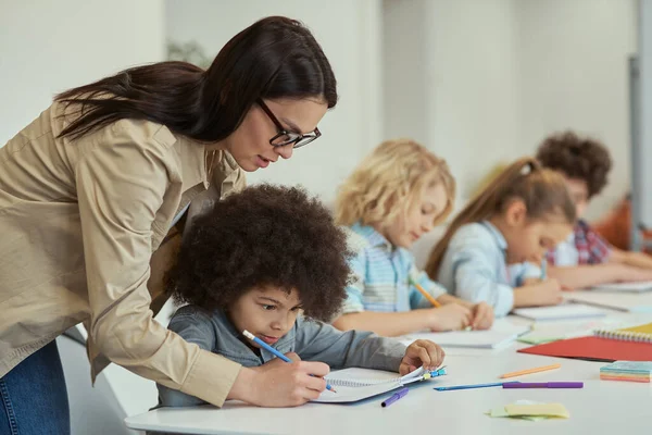 Profesora atenta y joven ayudando a un colegial. Niños sentados a la mesa, estudiando en la escuela primaria — Foto de Stock