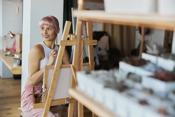 Model wanita muda positif dengan rambut merah muda duduk di kayu easel dengan kanvas — Stok Foto