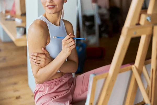 Smiling female model with graphite pencil sits at wooden easel in studio — Stock Photo, Image