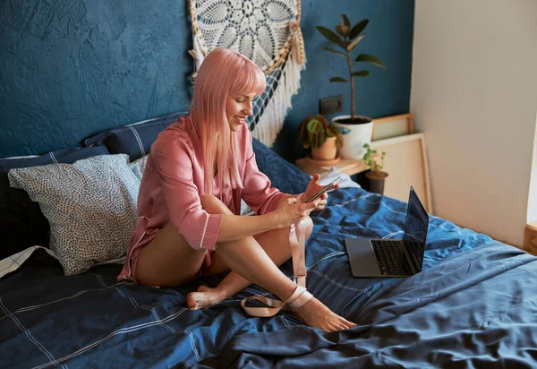 Aantrekkelijke vrouw surft internet met smartphone zitten in de buurt laptop op bed — Stockfoto