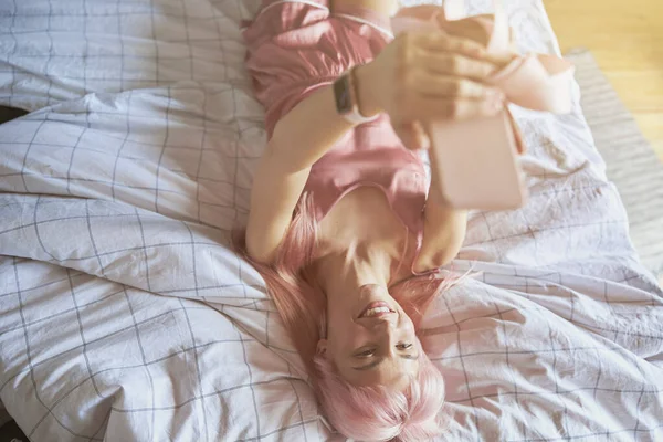 Glimlachende roze harige vrouw in elegante pyjama neemt selfie liggend op bed — Stockfoto