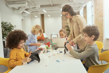 Teknoloji bilgini ol. Genç bayan öğretmenle STEM dersi sırasında çeşitli çocukların robot montajı ve programlaması