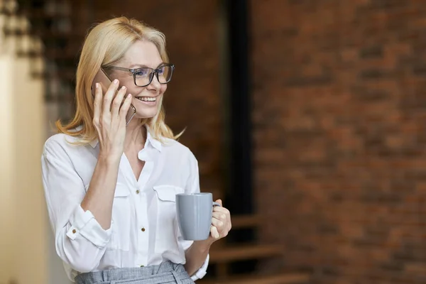 Sibuk setengah baya wanita dengan rambut pirang dan kacamata di kemeja putih minum kopi atau teh sambil berbicara di telepon, berdiri di rumah — Stok Foto