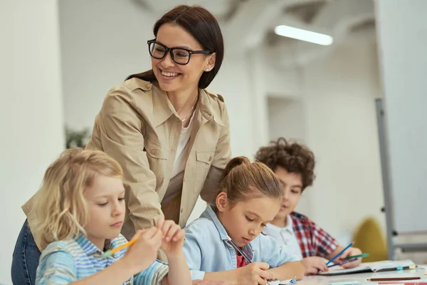 Glad ung kvinnlig lärare i glasögon hjälper sina små elever i ett klassrum. Barn som sitter vid bordet och studerar i grundskolan — Stockfoto